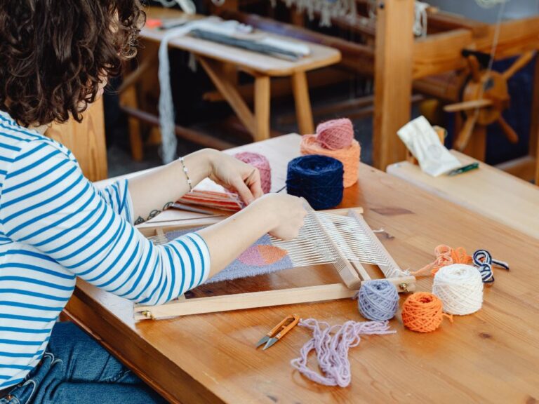 Pessoa produzindo artesanato em tear manual com fios coloridos sobre mesa de madeira, criando peça têxtil para venda.